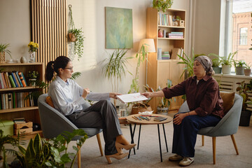 Middle aged Caucasian female psychologist sitting in modern office handing psychological test results to middle aged woman client wearing headscarf during therapy session © Seventyfour