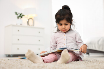 Fototapeta premium Cute little girl reading book on floor at home. Space for text