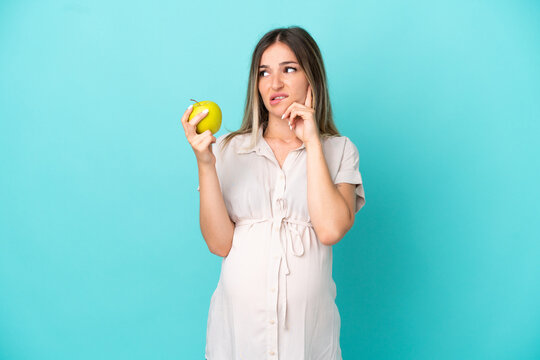 Young Romanian woman isolated on blue background pregnant and holding an apple while thinking