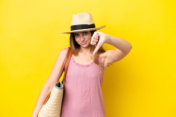 Fototapeta premium Young caucasian woman holding a beach bag isolated on yellow background showing thumb down with negative expression