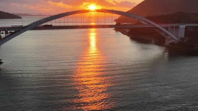 Cinematic golden hour at Omishima Bridge, sun peeking through the silver arch, 4K aerial.
