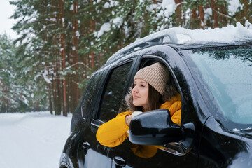Young woman in yellow winter jacket leaning from passenger side window of car during winter road trip through snowy forest enjoying calm landscape and travel atmosphere