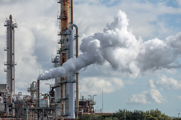 Industrial refinery plant complex emitting dense vapor cloud trail into partially overcast sky from rust-streaked refinery chimney, highlighting emissions, energy production and environmental impact. © thomasgweber