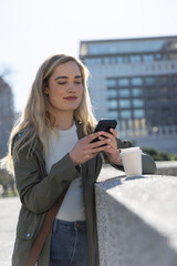 Fototapeta premium Woman in green field jacket leaning on concrete ledge in plaza, using smartphone with coffee cup