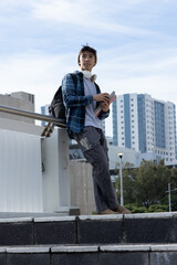 Asian teenage student standing on campus steps, leaning on handrail, holding notebooks and backpack
