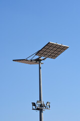 A tall metal pole equipped with multiple large LED floodlight panels and smaller spotlights stands under a clear blue sky during the day