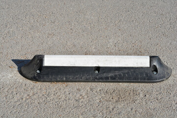 A black rubber parking wheel stop with a white reflective strip is bolted to the grey asphalt ground in an outdoor parking lot during the day