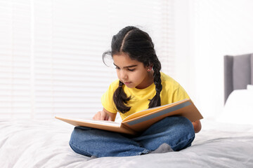 Fototapeta premium Cute little girl reading book on bed at home