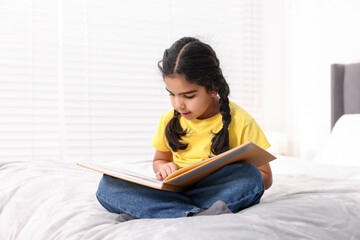Fototapeta premium Cute little girl reading book on bed at home. Space for text