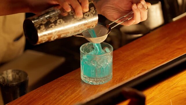 A professional bartender uses a Hawthorne shaker and a fine mesh strainer to double-strain a vibrant blue cocktail into a crystal rocks glass filled with ice at a trendy bar.