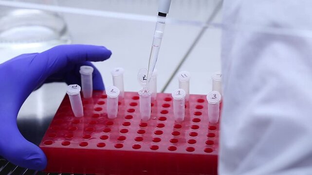 A close-up shot from behind a scientist in a white coat shows the scientist adding clear liquid to a small eppendorf tube in a red laboratory stand using an automatic pipette.