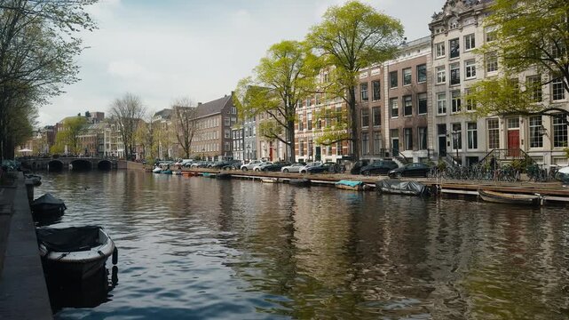 Singel canal in Amsterdam, showing historic Dutch houses, moored boats, and trees reflecting in the water on a sunny spring day