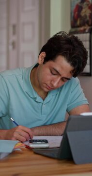 College student studies at home, writing notes beside a calculator and tablet while preparing for math and science exams, with subtle lateral movement, vertical format.