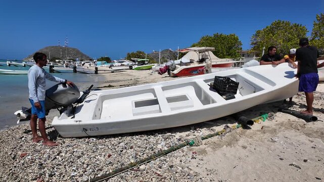 Coastal scene of local men preparing small motorboat on sandy Caribbean beach in Los Roques Venezuela. Coastal livelihood, Fishing boats and anchored vessels are visible in background