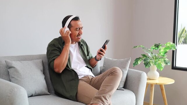 Happy carefree Asian man in stylish headphones listening to music from smartphone moving head, hand to rhythm, singing sing aloud