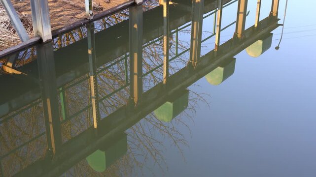 Reflection of an iron sluice gate and surrounding pond embankment on the calm surface of a quiet fishpond, with gentle ripples forming as water flows softly through the grating in the background