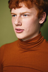 Fototapeta premium Portrait of young man with red hair and blue eyes wearing orange turtleneck sweater, looking sideways on green background with serious expression and clean skin in studio light.