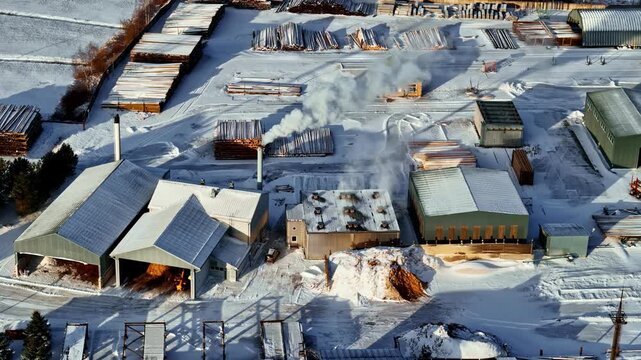 Winter view of sawmill buildings with smoking chimney and stacked timber across a snow covered yard