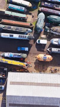 Top-down vertical drone over Stanstead Abbots marina in Hertfordshire, UK, flying forward above main building and boats on hardstanding, with River Lea to the right in daylight.