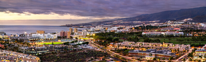 Fototapeta premium Scenic panoramic twilight view of the illuminated city streets, stadium, and seaside resorts of Playa de las Americas against a mountainous backdrop in Tenerife, Spain