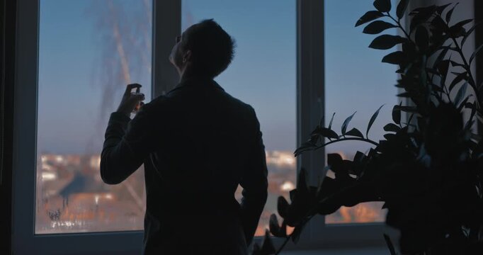 A man sprays perfume while standing by a window in a classic suit