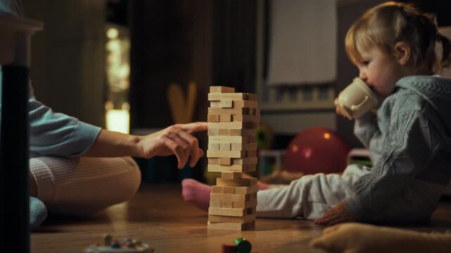 A mother and young daughter engage in a delightful evening of bonding on the floor, constructing an unstable tower with wooden Jenga blocks, promoting learning, entertainment