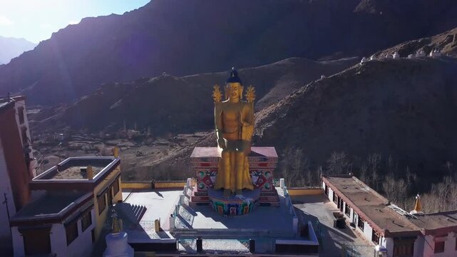 Cinematic drone pullback from a close-up of the golden Maitreya Buddha statue at Likir Monastery revealing the whitewashed monastery complex and barren Himalayan valley in winter sunlight