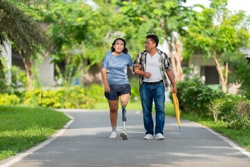 Obraz premium Happiness couple walking on pathway in park woman wearing prosthetic leg and support man holding umbrella. Smiling disabled female with artificial limb strolling with her partner in outdoors.