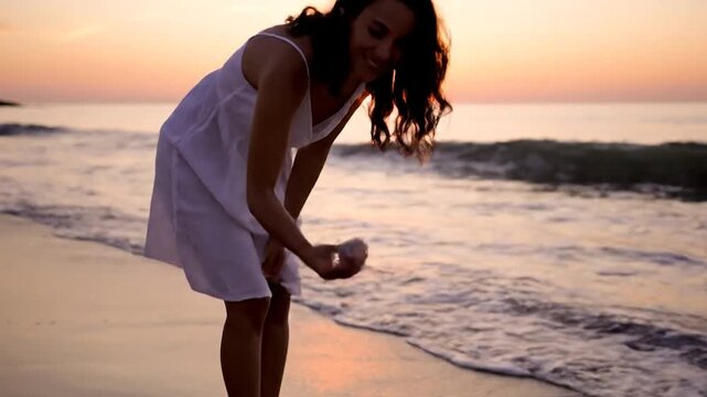 Woman in White Dress Finds Seashell on Beach at Sunset.