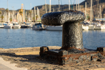 Fototapeta premium A weathered, rusted bollard stands prominently in the foreground of harbor of Cartagena, with numerous sailboats and yachts moored in the background under a clear sky
