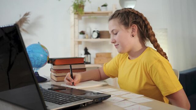 Girl study at desk writing flashcard and homework with laptop and book child student focuses on learning and education holding card and pen beside globe and stack of book in tidy study area smile