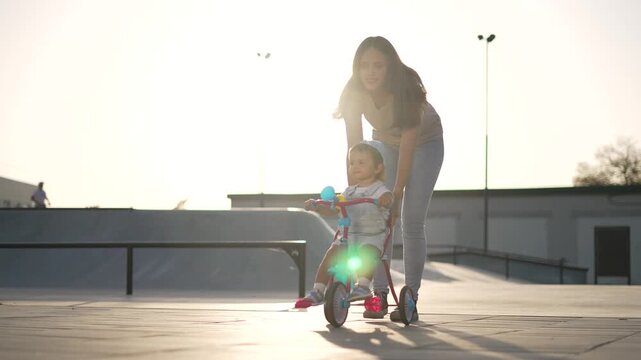 Mother pushes toddler on tricycle at urban park skate area in warm sunlight with parent guiding child for balance training and learning while caregiver holds handlebar supporting steady ride
