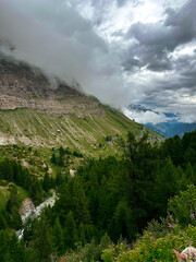 Lush green slopes and forests cover the lower mountainsides, contrasting with the grey rock and white snow of the high altitudes. Patches of blue sky are visible through the cloud cover, evoking a sen © Марина Сергеева