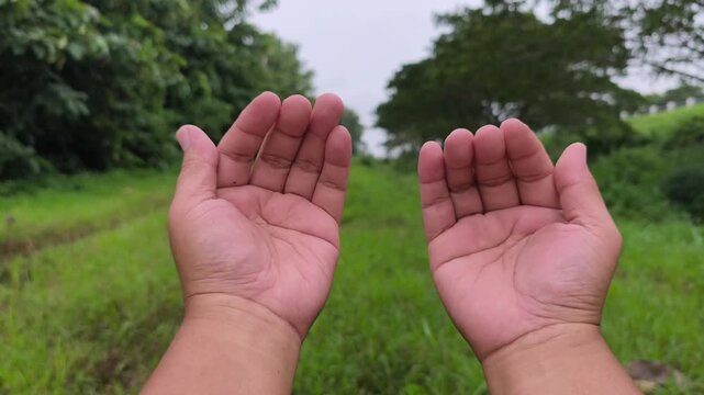 Open palms raised in prayer against lush green field under cloudy sky background