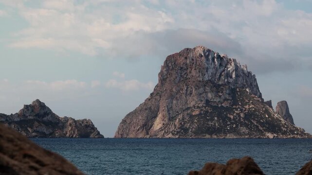 Static timelapse of Es Vedra in Ibiza with fast moving clouds and boats crossing turquoise Mediterranean waters.