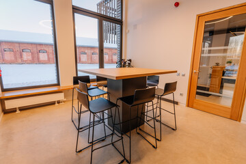 Fototapeta premium Modern interior of a minimalist office kitchen featuring a large wooden table surrounded by black metal stools and large windows with natural light
