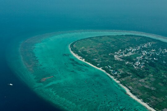 Wide aerial shot of Thoddoo island showcasing the local village, dense palm forests, and an expansive turquoise coral reef surrounding the coastline in the Maldives.