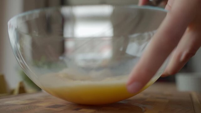 Hands whisk beaten eggs in glass bowl on wooden kitchen counter with cracked eggshells. Person preparing breakfast. Concept of home cooking food preparation and recipe ingredients.