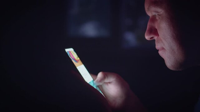 A man's face against the backdrop of a window at night, browsing apps on his mobile phone, side view.