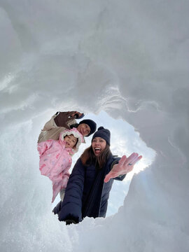 Joyful three-generation Asian family selfie portrait in Japan, featuring grandparents, parents, and a child smiling together through a snow hole, capturing a perfect winter vacation moment.
