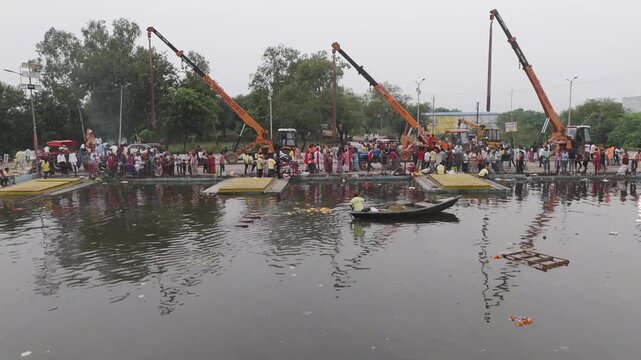 Durga idol immersion by crane at riverside kund, Festival crowd at Durga visarjan ghat, Crane lifting goddess idol for immersion ritual, Devotees gathering for Durga visarjan near water kund.