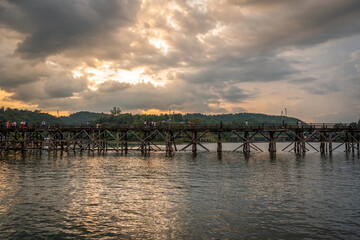 Obraz premium Dramatic sunset over wooden Mon Bridge with silhouette traveler crossing calm river in Sangkhlaburi, Kanchanaburi, Thailand