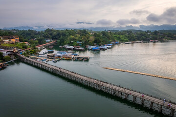Obraz premium Aerial panoramic view of Mon Bridge in Sangkhlaburi, Kanchanaburi, Thailand stretching across calm river beside floating house and lush green hillside village under soft evening sky