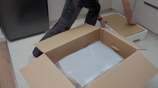 Person removes foam packaging from boxed built in appliance on kitchen floor. Concept of appliance installation preparation and home product unboxing process.