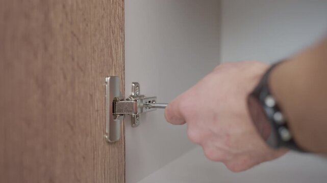Person adjusting metal cabinet door hinge using screwdriver inside wooden kitchen cabinet. Concept of home repair furniture installation and maintenance.