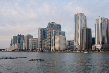 Fototapeta premium 10.12.2025 Sharjah, UAE. Sunset skyline of Sharjah with modern skyscrapers and luxury residential towers along the Corniche waterfront under a golden sky, United Arab Emirates.