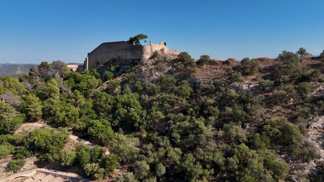 Aerial drone flying backward and rising from the hillside to reveal the stone walls of Xativa Castle and the surrounding Valencia landscape.