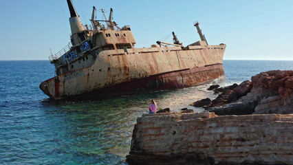 Young Caucasian woman sitting on coastal rocks, looking at the rusty Edro III shipwreck partially submerged in the clear turquoise waters of the Mediterranean Sea near Paphos, Cyprus