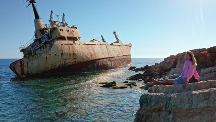 Fototapeta premium Young woman sitting on a rocky seashore admiring the iconic Edro III shipwreck, a popular tourist landmark near Paphos, under a bright blue sky on a clear day