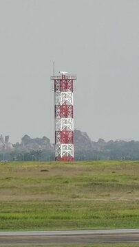 Tall red and white lattice communication tower with antennas and equipment on top, located in a grassy field beside a paved runway. Mountainous rocky terrain forms the natural backdrop.